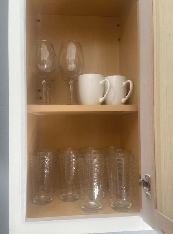 A wooden cabinet with two shelves; the top shelf holds two wine glasses and two white mugs, and the bottom shelf contains several clear plastic cups.
