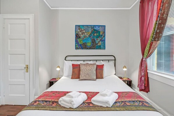 A neatly made bed with a colorful bedspread, two bedside lamps, a painting above the bed, and a window with curtains.