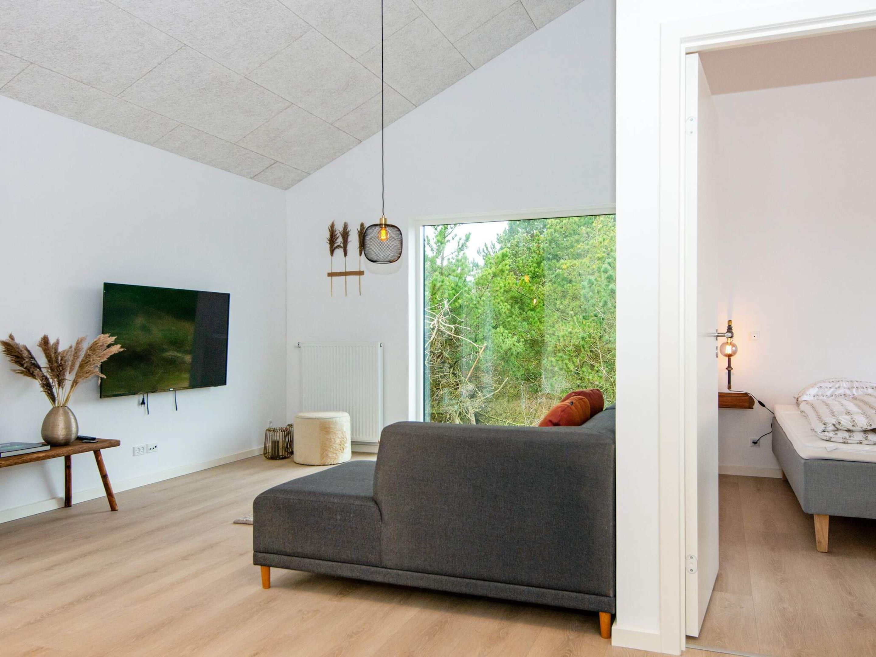 Ein modernes Wohnzimmer mit einem grauen Sofa, einem Flachbildfernseher und Blick auf Grünflächen.