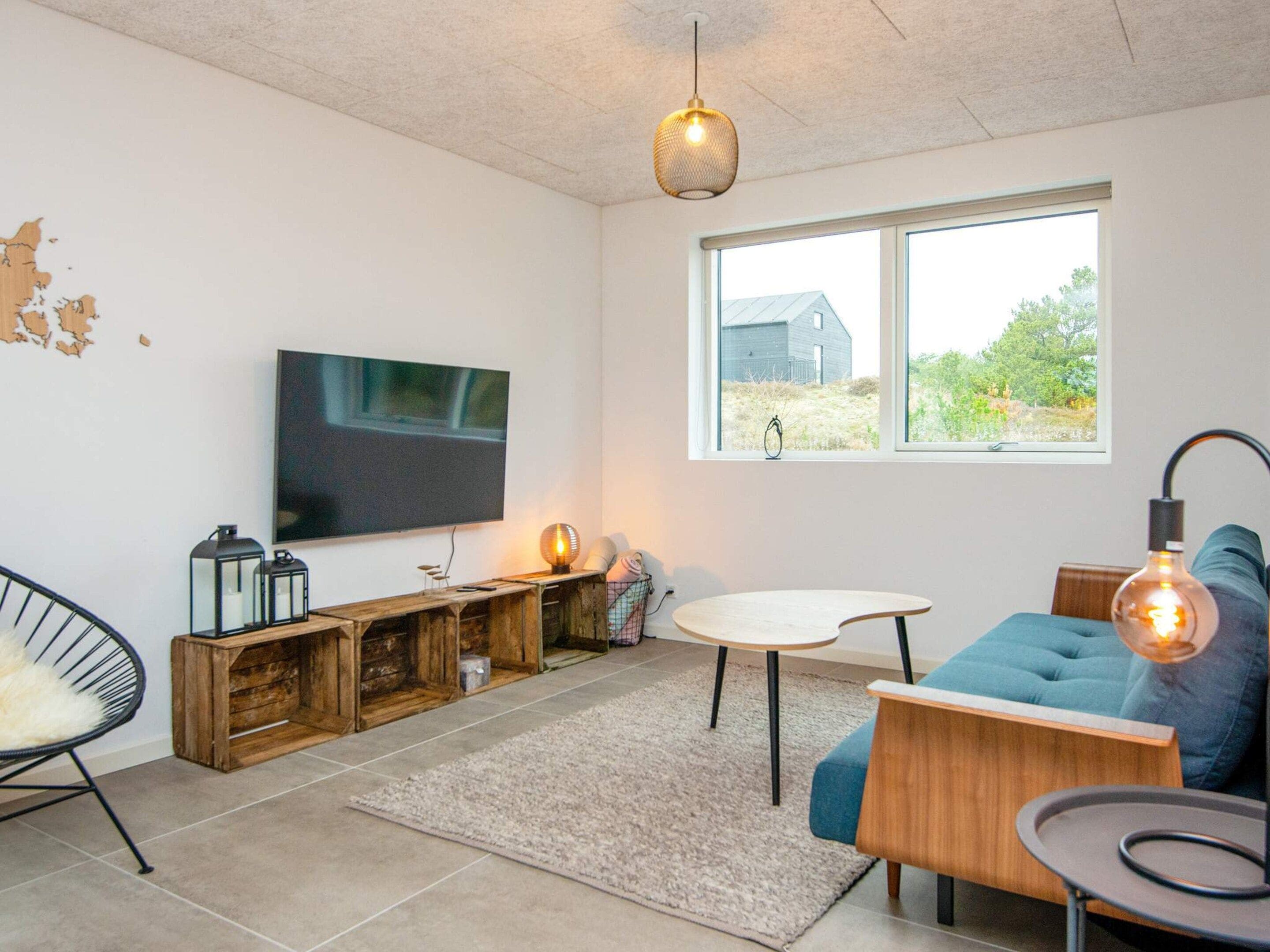 A modern living room with a flat-screen TV, wooden storage, a blue sofa, and a large window with a view of greenery.