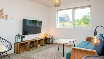 A modern living room with a flat-screen TV, wooden storage, a blue sofa, and a large window with a view of greenery.