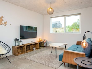 A modern living room with a flat-screen TV, wooden storage, a blue sofa, and a large window with a view of greenery.