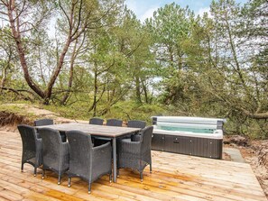 Um deck externo com mesa de madeira e cadeiras de vime, cercado por árvores e uma banheira de hidromassagem.