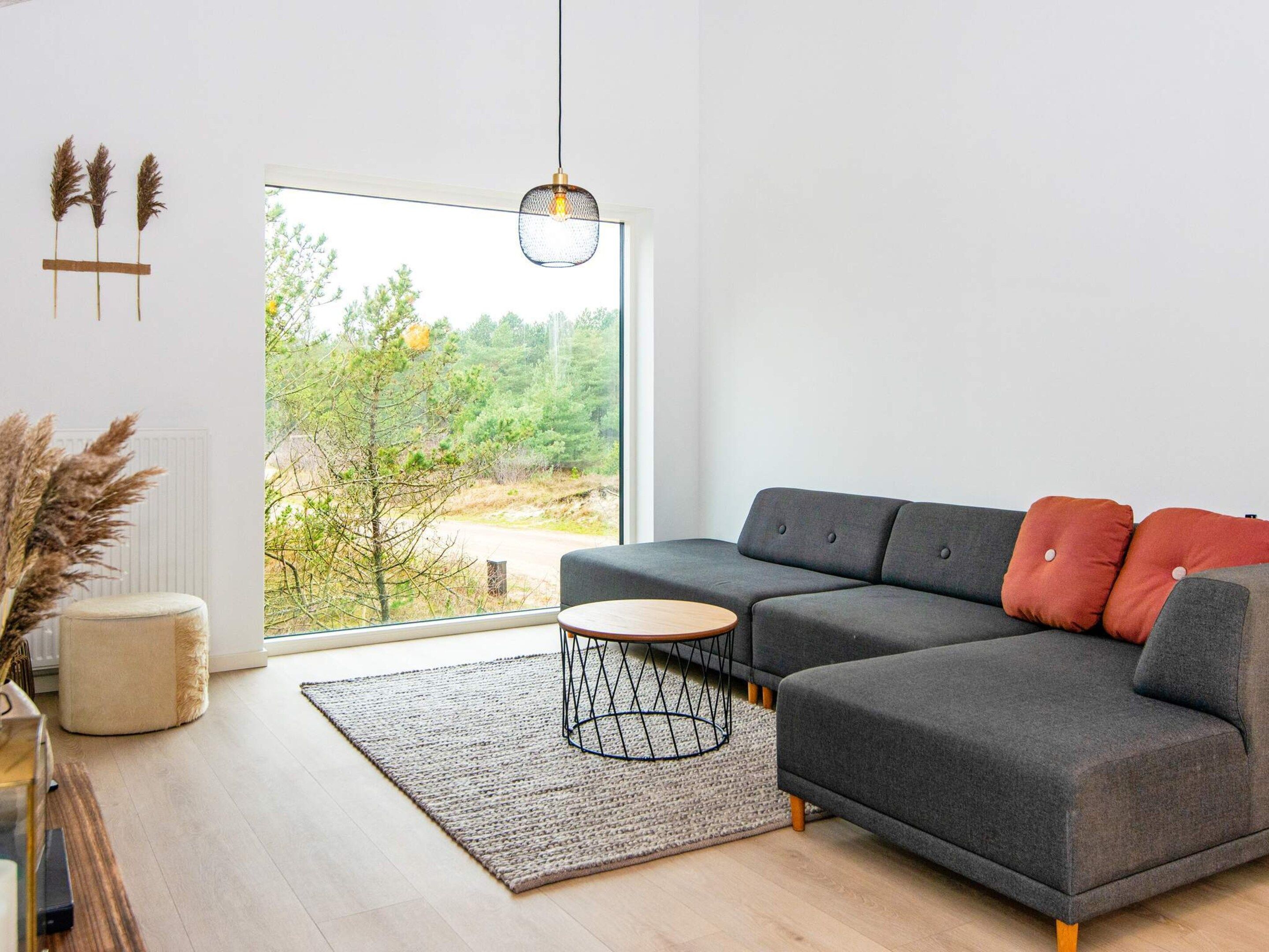 A modern living room with a grey sectional sofa, a round coffee table, and a view of a forest through large windows.