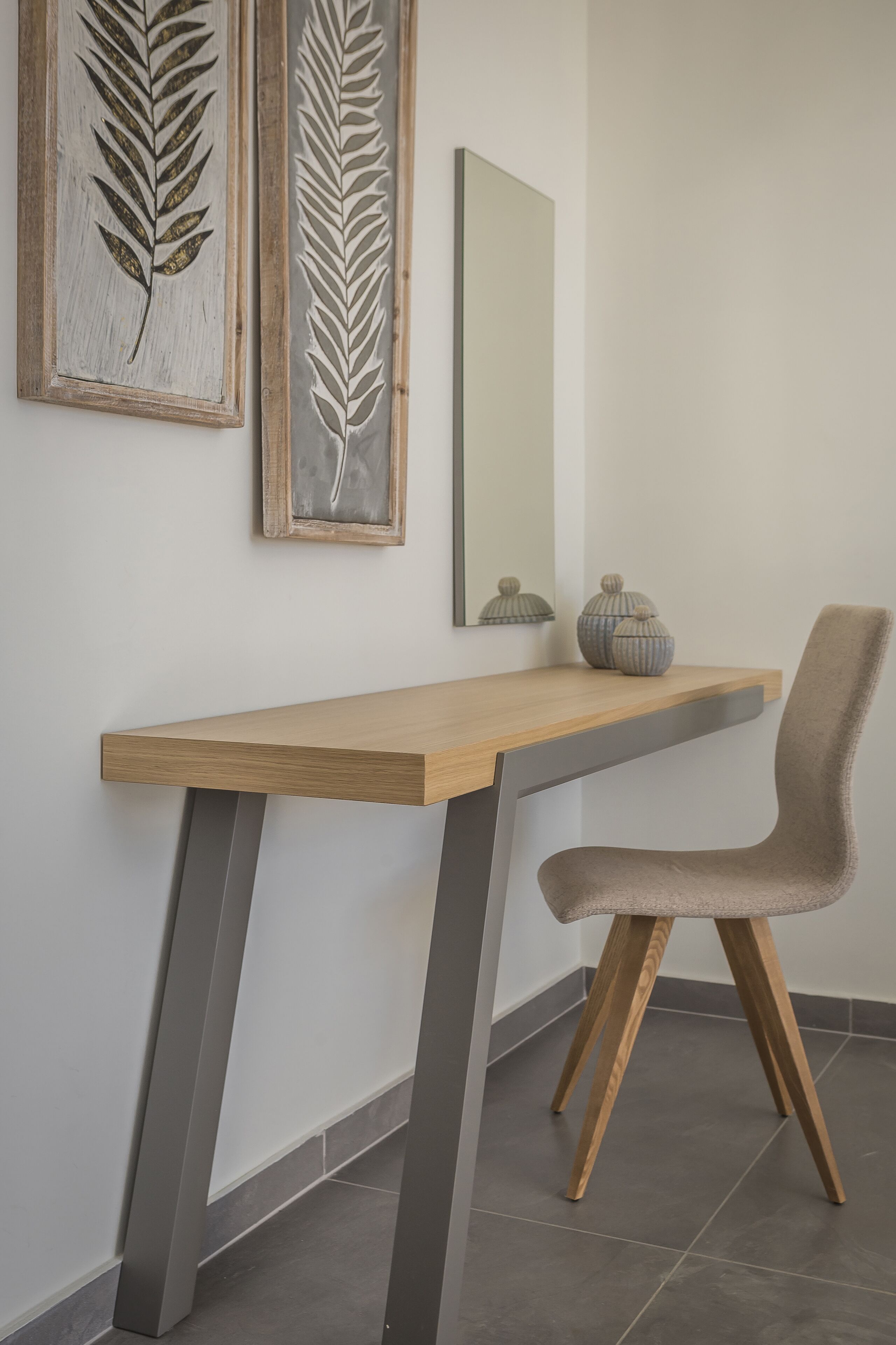 A modern desk with a chair, a mirror, and two framed leaf designs on the wall.