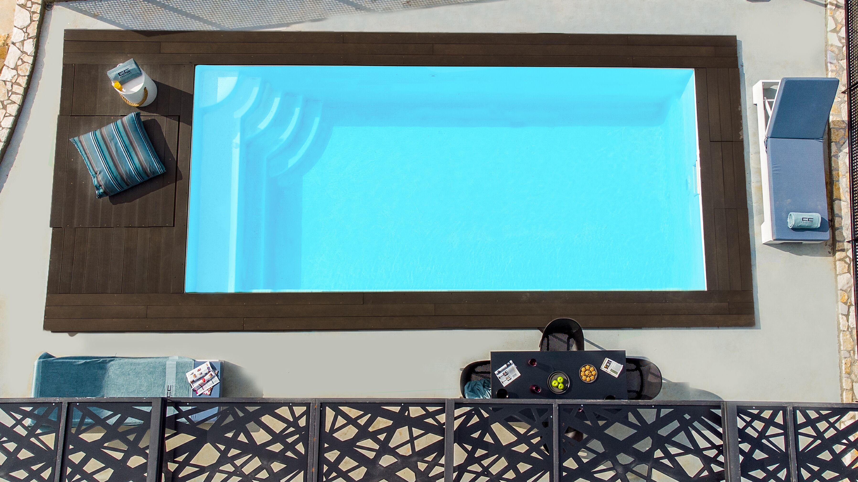 A rectangular pool with a dark wooden border, a lounge chair, a towel, and a small table with a bottle and a bowl of fruit.