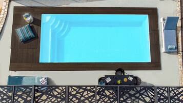 A rectangular pool with a dark wooden border, a lounge chair, a towel, and a small table with a bottle and a bowl of fruit.