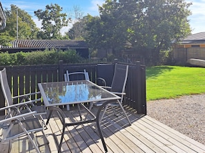 A deck with a glass table and chairs, surrounded by a wooden fence and a grassy lawn. - 6 Person Holiday Home in Hadsund (Hadsund)