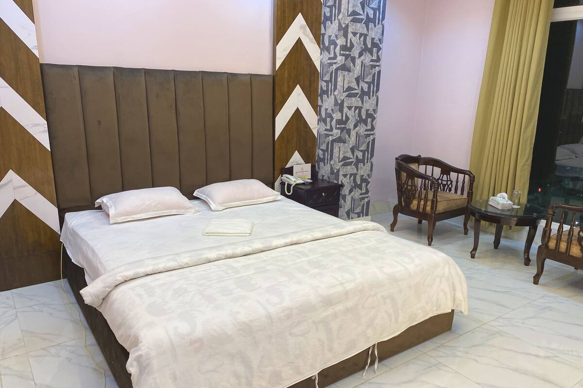A neatly made bed with pillows and a folded blanket, a bedside table with a lamp, a chair, and a patterned curtain.