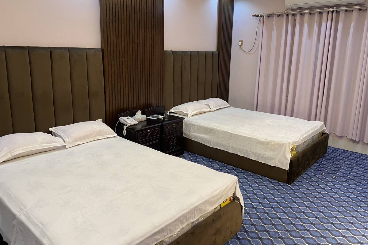 A hotel room with two beds, a wooden headboard, a bedside table with a phone and tissues, and a window with curtains.