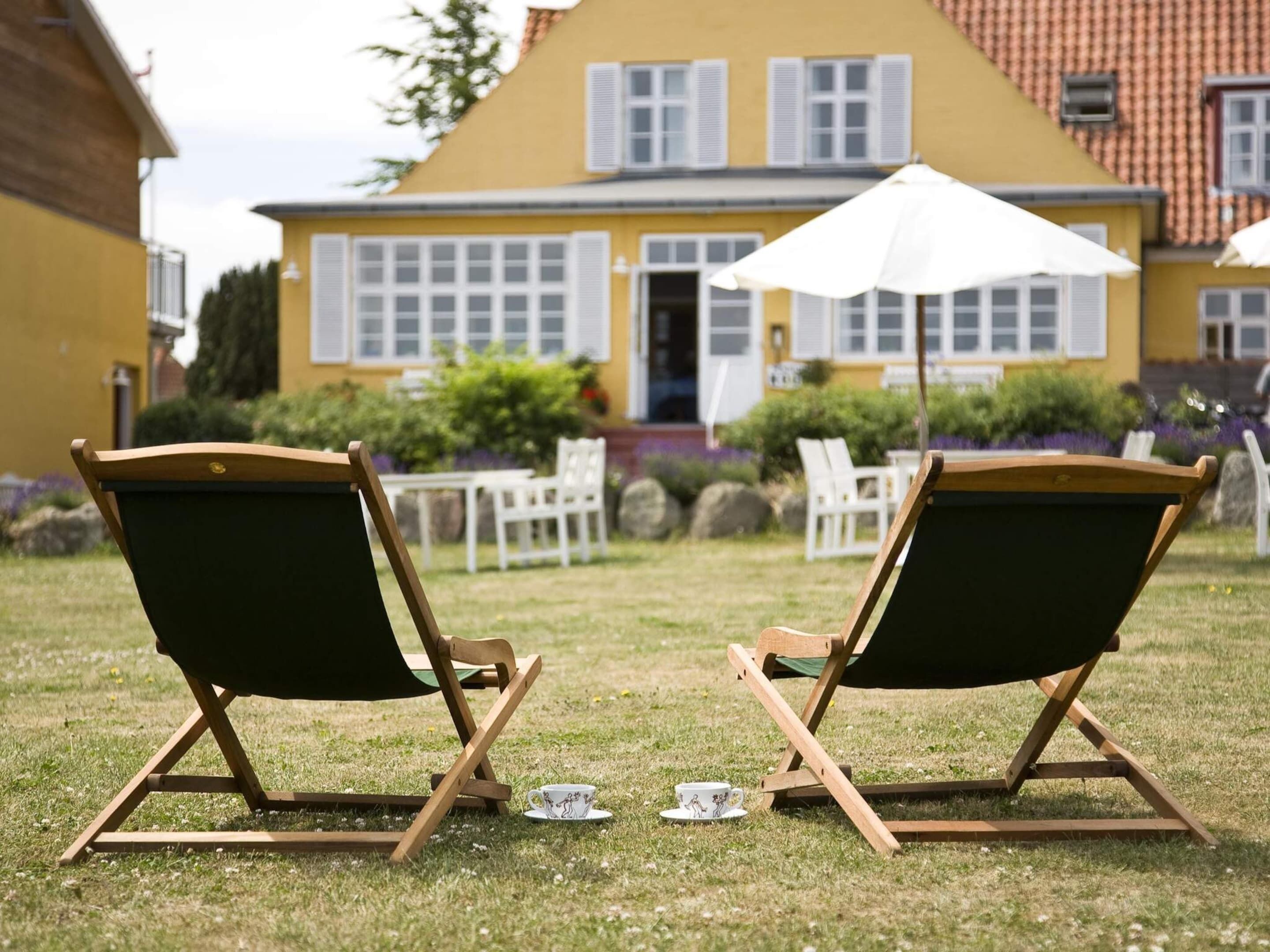 Two wooden lounge chairs with green cushions in a grassy area in front of a yellow building with white shutters.