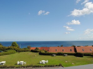 A coastal view with a building featuring a red roof, a grassy lawn with outdoor seating, and the ocean in the background. - 2 Person Holiday Home in Gudhjem (Gudhjem)