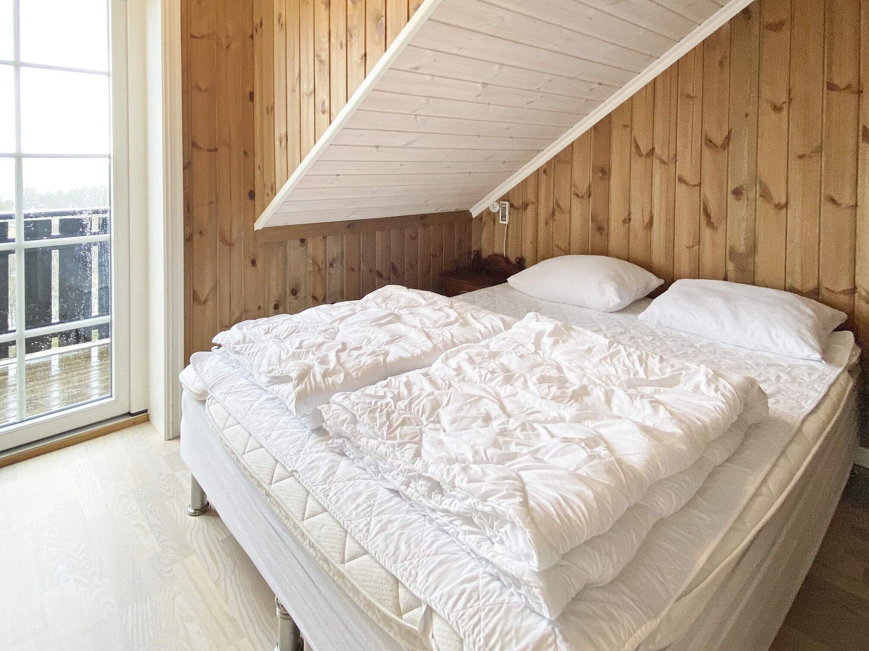 A bedroom with a sloped ceiling and wooden walls, featuring a large bed with white bedding and a window providing a view.
