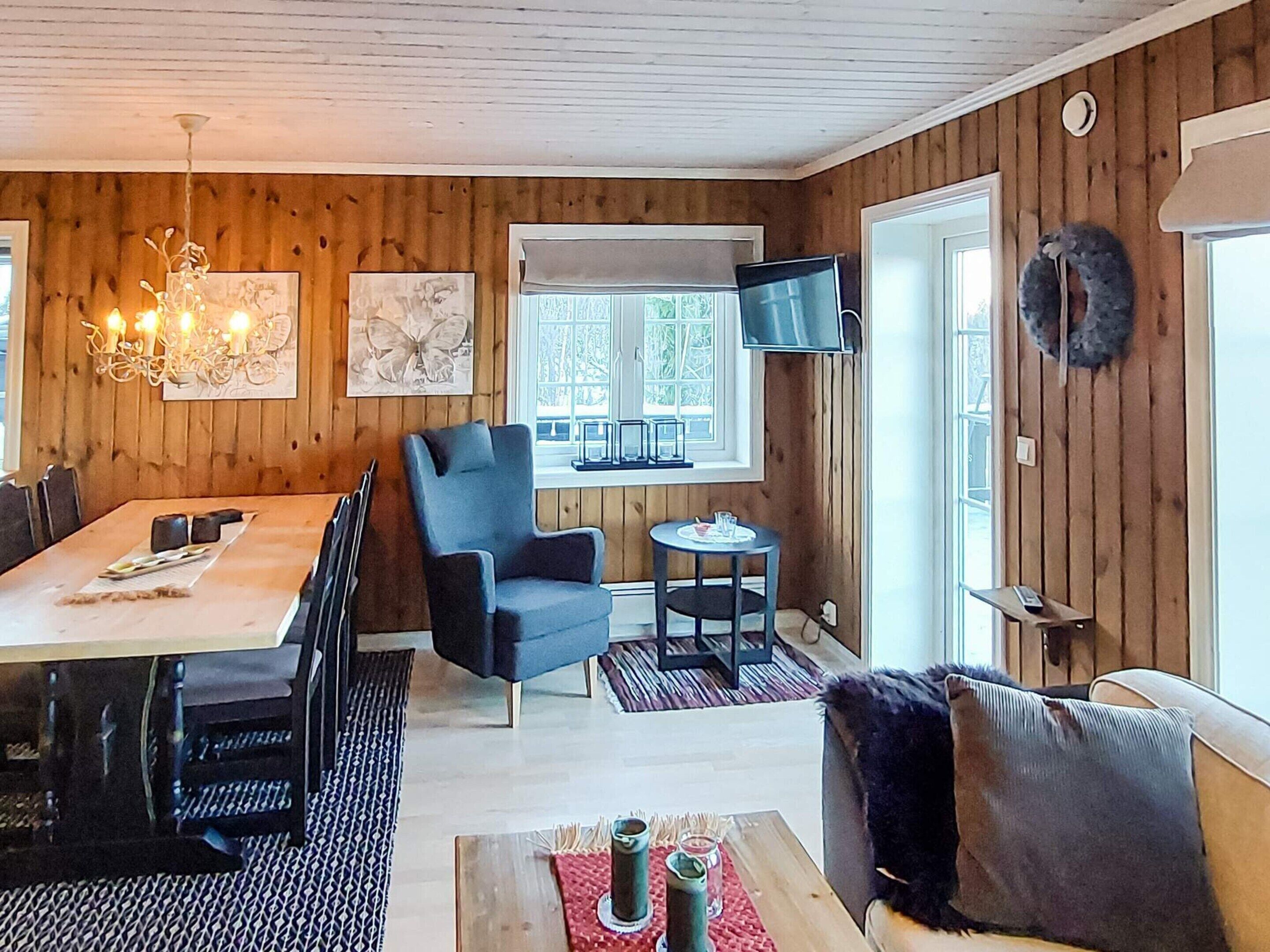 A cozy room with a wooden interior, a dining table with chairs, a blue armchair, a small round table with a chair, a sofa, and a TV mounted on the wall.