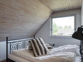 Un dormitorio con techo inclinado, una ventana que ofrece vistas a árboles y edificios, una cama con ropa de cama blanca y almohadas estampadas, y una lámpara de escritorio negra.