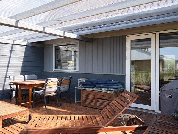 A covered patio area with a wooden table, chairs, a lounge chair, and a sliding glass door leading to a garden.