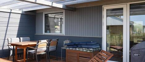 A covered patio with a wooden table, chairs, a lounge chair, and a grill area.