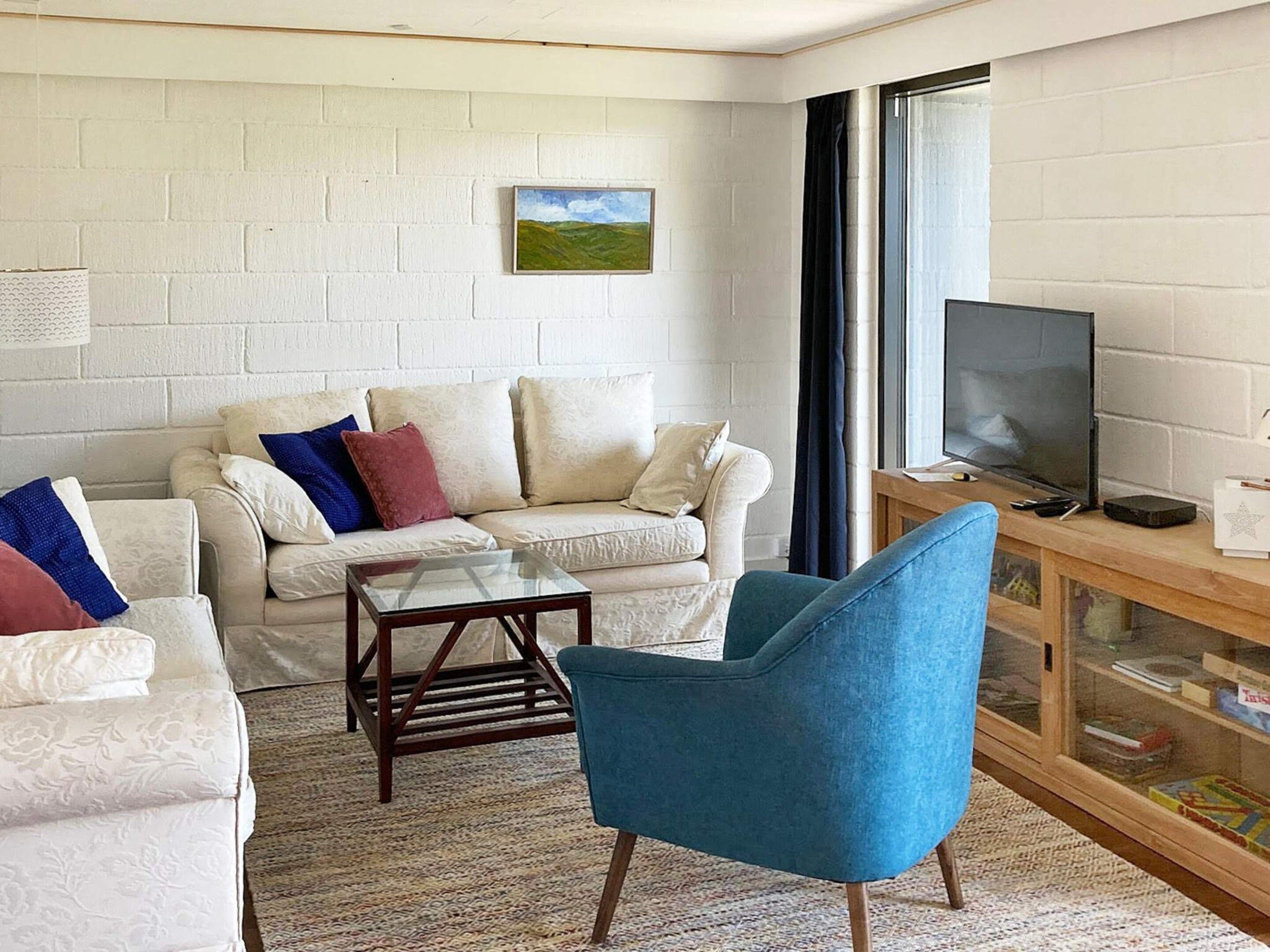 A living room with a white brick wall, a sofa with cushions, a blue armchair, a glass coffee table, and a television on a wooden stand.