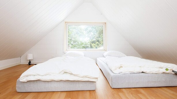 A small, white-walled room with a sloped ceiling and a window. There are two beds with white bedding and a lamp on a wooden floor. - 6 Person Holiday Home in Kolind (Kolind)
