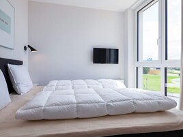 Ein modernes Schlafzimmer mit einem großen Bett, einem an der Wand befestigten Fernseher und einem Fenster mit Blick ins Freie.