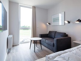 Una habitación de hotel moderna con un sofá gris, una pequeña mesa redonda, una cama y un amplio ventanal con cortinas.