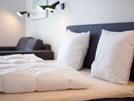 Una cama con ropa de cama blanca y dos almohadas, un cabecero oscuro y una lámpara fijada a la pared.