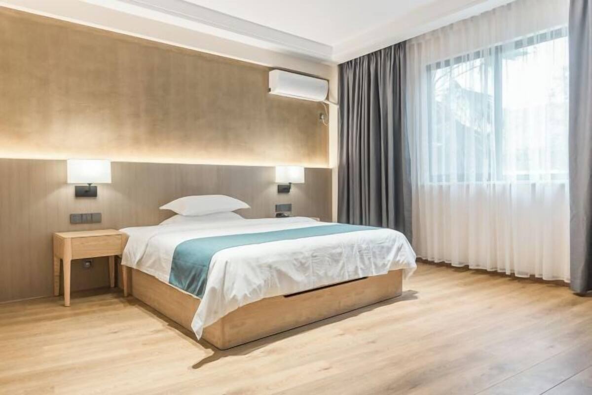 Moderni hotellihuone, jossa on puinen sänky, kaksi yöpöytävalaisinta, suuri ikkuna verhoilla sekä seinälle asennettu ilmastointilaite.
