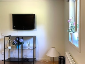 A room with a mounted television, a black console table with decorative items, a standing lamp, and a window with a potted plant. - 4 Person Holiday Home in Uddevalla (Uddevalla)