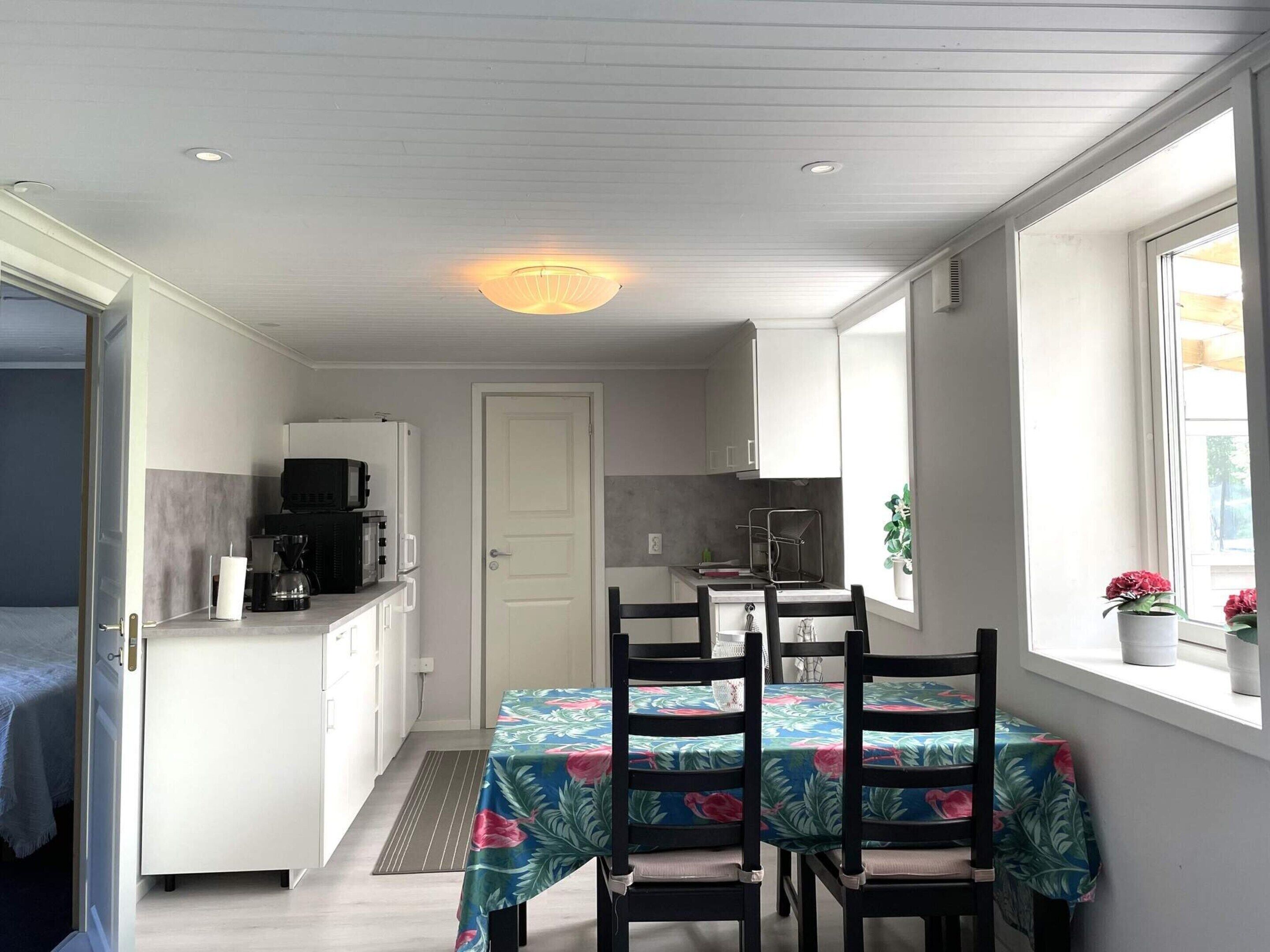 A compact kitchen with white cabinets, a microwave, and a coffee maker. There is a dining table with chairs covered in a floral tablecloth.