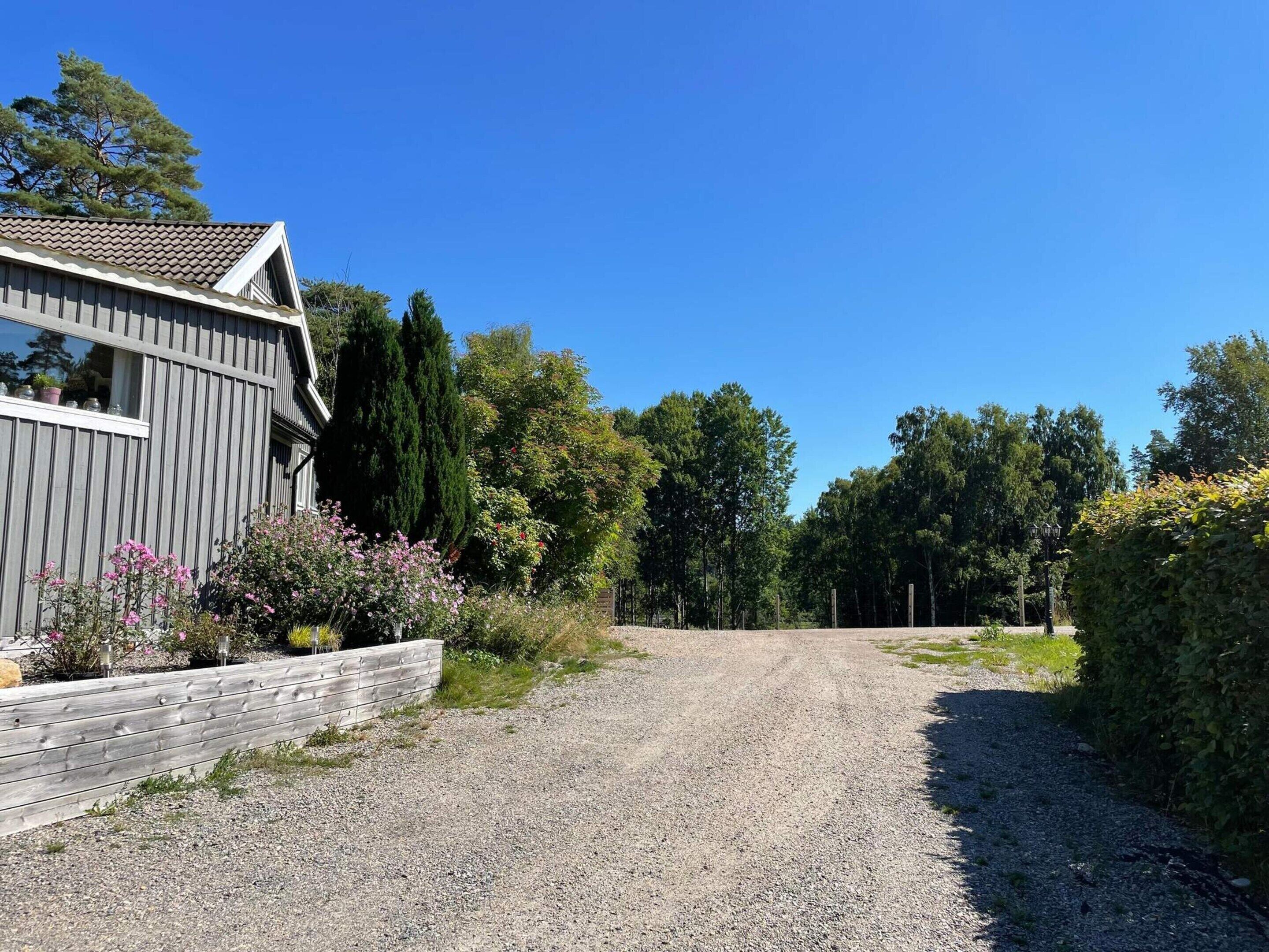 Une allée de gravier mène à une maison en bois dotée d’un toit en pente, entourée d’arbres et d’arbustes.