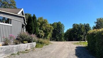 A gravel driveway leading to a wooden house with a sloped roof, surrounded by trees and shrubs.