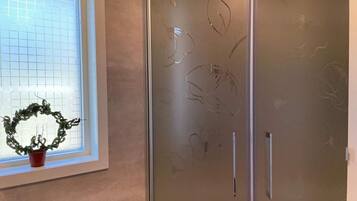 A bathroom with a frosted glass shower door, a window with a grid pattern, and a potted plant on the windowsill.