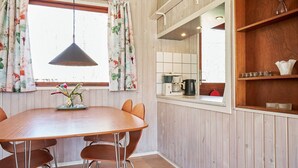 A dining area with a wooden table, chairs, a hanging light fixture, a window with curtains, and a kitchen area visible through an open door. - 4 Person Holiday Home in Aakirkeby (Akirkeby)