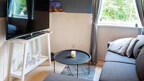 A living room with a flat-screen TV on a white stand, a round coffee table with a candle, a grey sofa, and a window with curtains. - 6 Person Holiday Home in Stromstad (Stromstad)