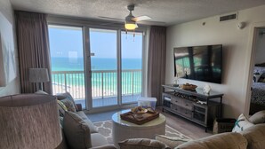 A living room with a large TV, a ceiling fan, and a view of the beach.