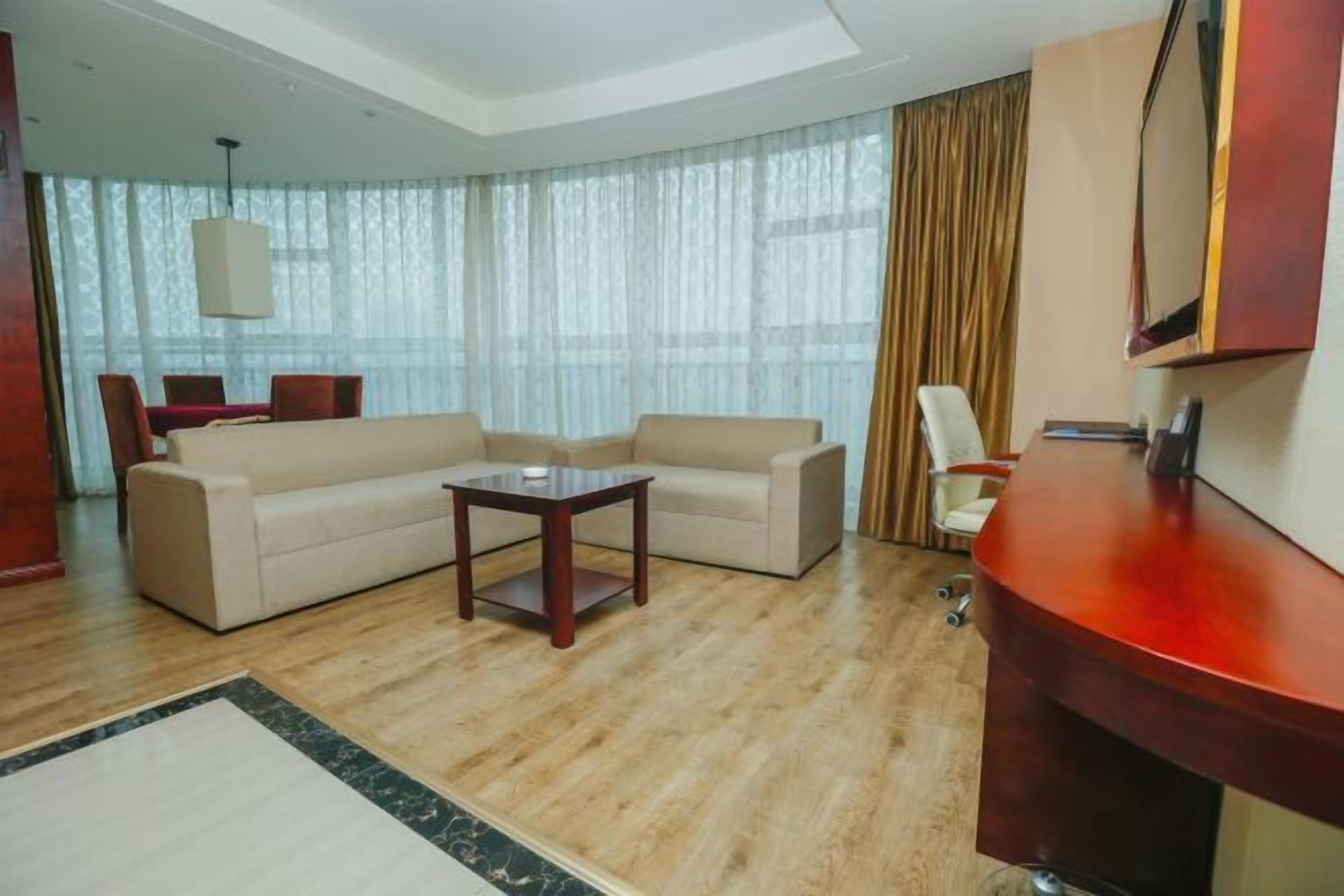 A modern hotel room with a wooden floor, a beige sofa, a small wooden table, a red accent wall, and a large window with sheer curtains.