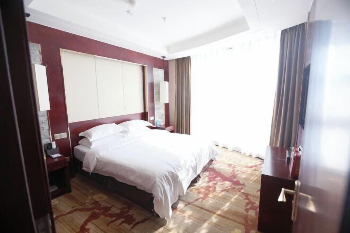 A hotel room with a large bed, a nightstand, a desk, a chair, a wardrobe, and a window with curtains.