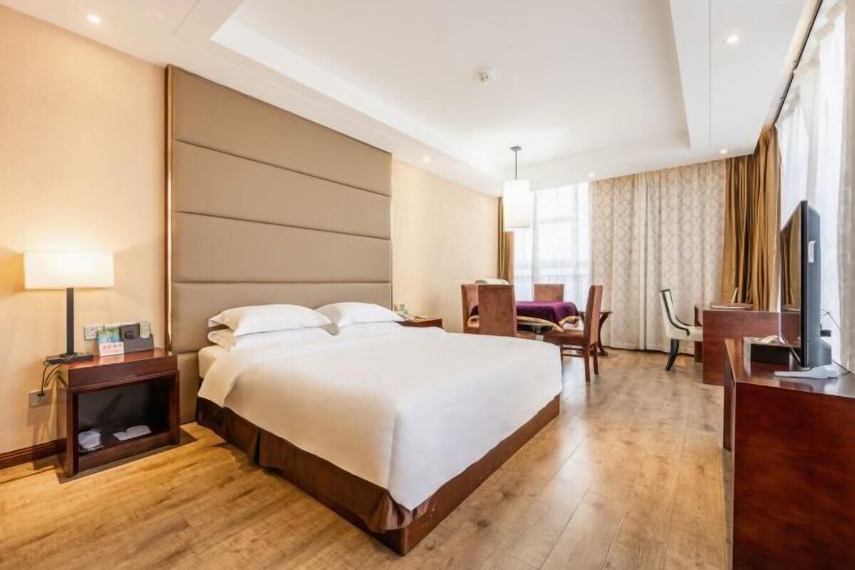 A hotel room with a large bed, a desk, a chair, a TV, and a lamp.