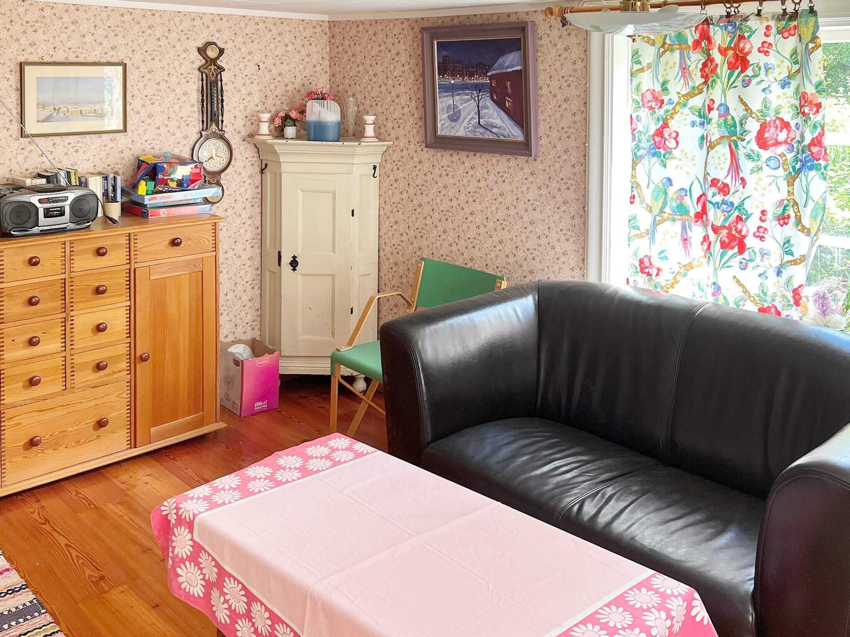 A living room with a black leather sofa, a wooden cabinet with drawers, a green chair, and a table with a pink floral tablecloth.