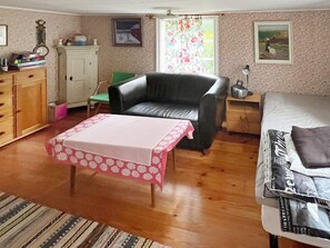A room with a black leather sofa, a wooden cabinet, a small table with a pink tablecloth, a bed with a blanket, and a window with floral curtains. - 7 Person Holiday Home in Algaras (Älgarås)