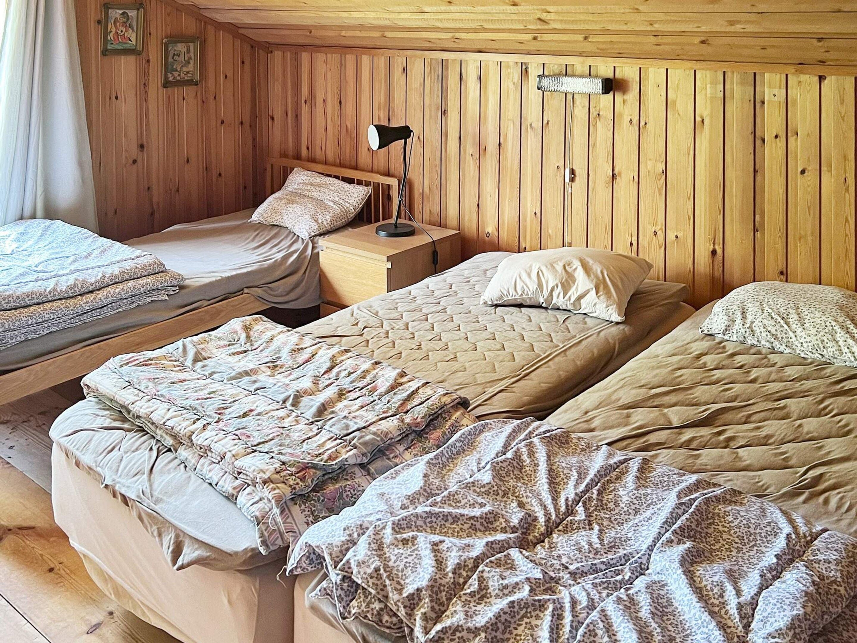 A room with three beds, wooden walls, and a nightstand with a lamp.