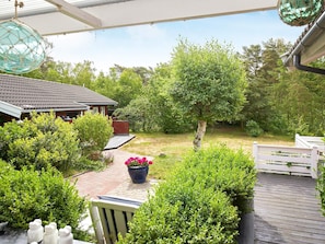 A covered outdoor area with a wooden deck, a potted plant with flowers, and a view of a house with a tiled roof.