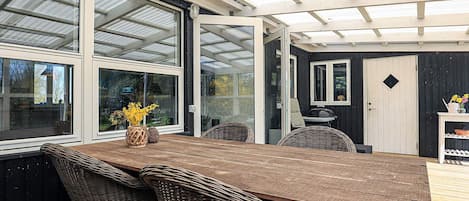 A covered outdoor dining area with a wooden table and wicker chairs, large windows, and a door.