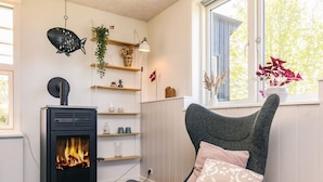 A room with a wood-burning stove, a chair with a cushion, shelves with decorative items, and a window with a view of trees. - 6 Person Holiday Home in Vinderup (Vinderup)