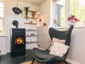 A room with a wood-burning stove, a chair with a cushion, shelves with decorative items, and a window with a view of trees. - 6 Person Holiday Home in Vinderup (Vinderup)