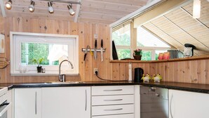 Cozinha moderna com armários brancos, lava-louças em aço inoxidável e janela com vista para o verde.