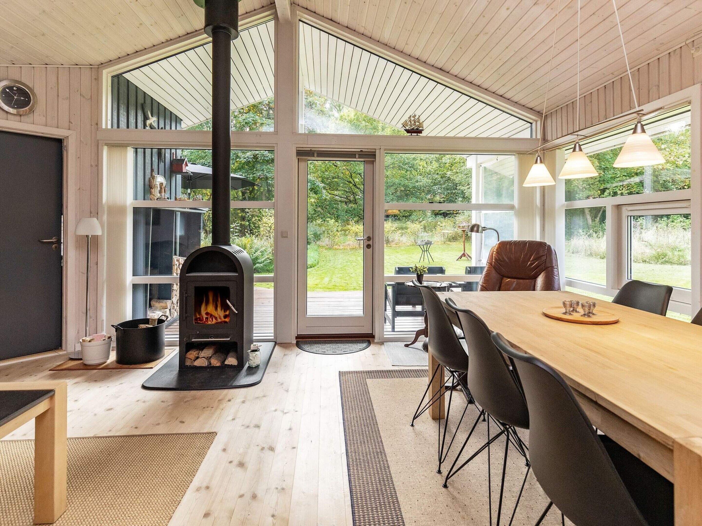 Un interior moderno con estufa de leña, comedor con sillas y amplias ventanas con vistas al exterior.