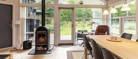 A modern interior with a wood-burning stove, a dining area with chairs, and large windows offering a view of the outdoors.