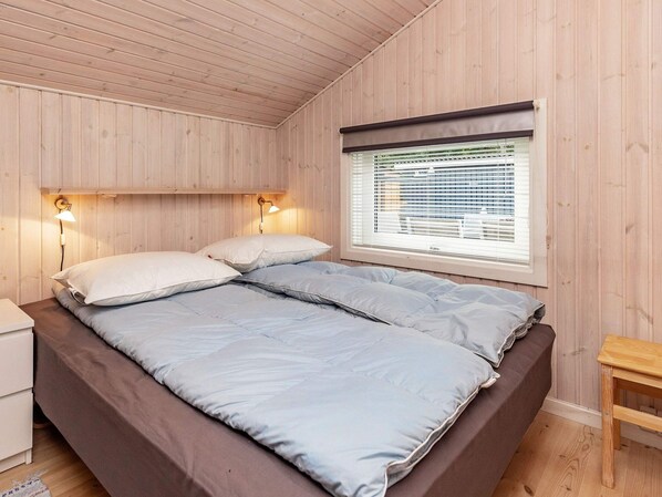 A single bed with two pillows, a bedside table, a nightstand, a window with blinds, and a wooden bench.