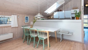 Uma sala de jantar com uma mesa branca e cadeiras verdes, uma cozinha com armários e uma luminária pendente, e uma janela com cortina.